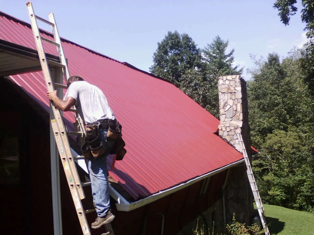 Roofer on a ladder beside a red metal roof and stone chimney — Soffit & Fascia Repair by Roofing Brothers in Waynesboro